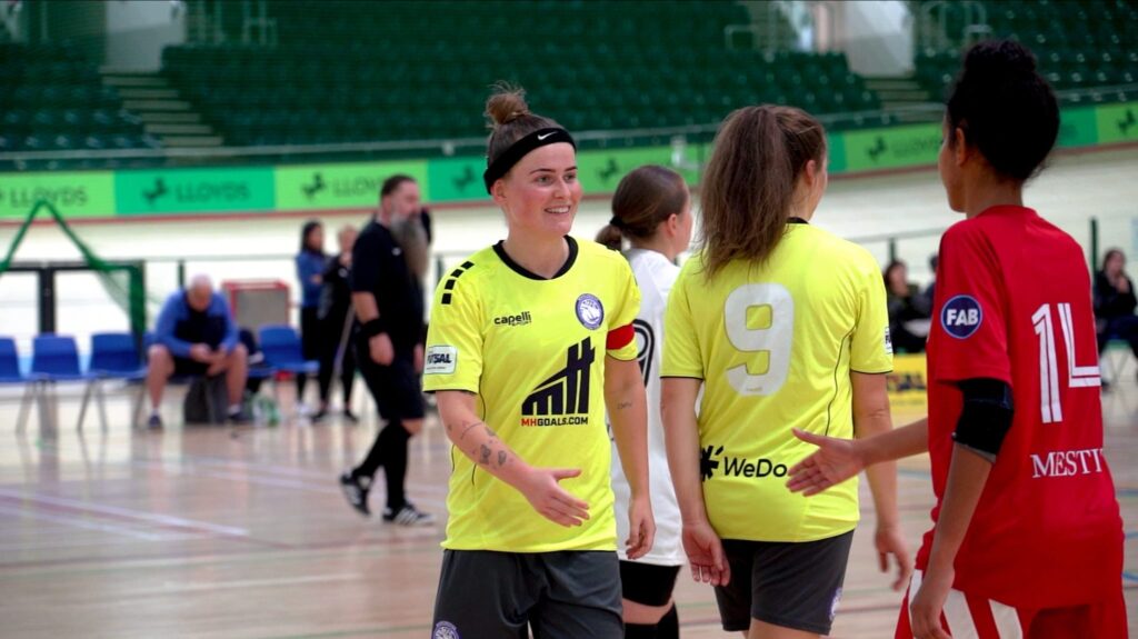 Women futsal players shaking hands after a match, in an indoor futsal setting.